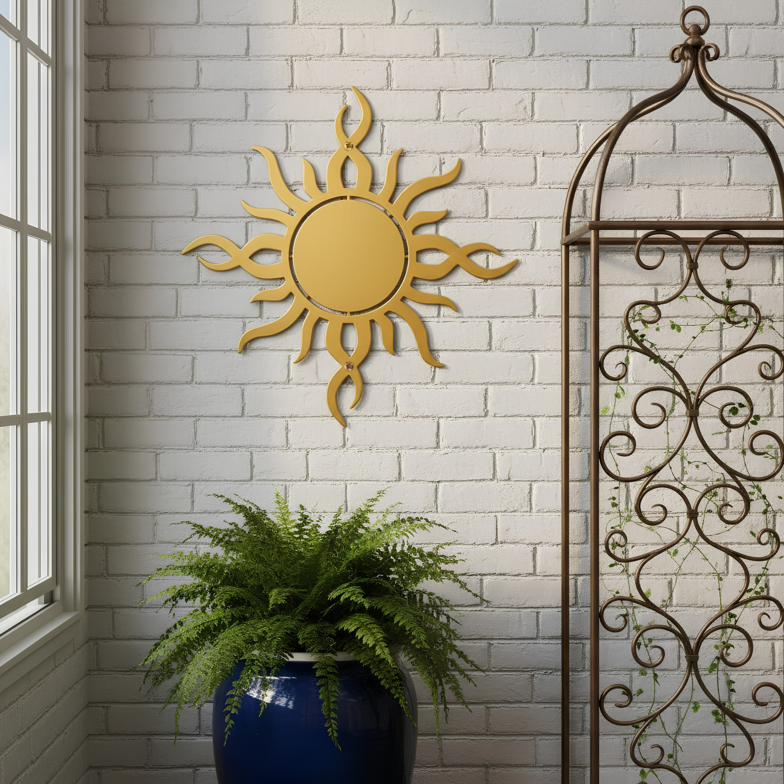 A gold-colored metallic sunburst sign hung on a white brick interior wall, positioned between a large window and a decorative brown iron trellis above a potted green fern.