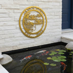 A gold-colored circular metal sign with a koi fish motif reading "Jane's Pond Est. 2022," mounted on a light stacked-stone wall directly above a backyard pond with swimming koi.