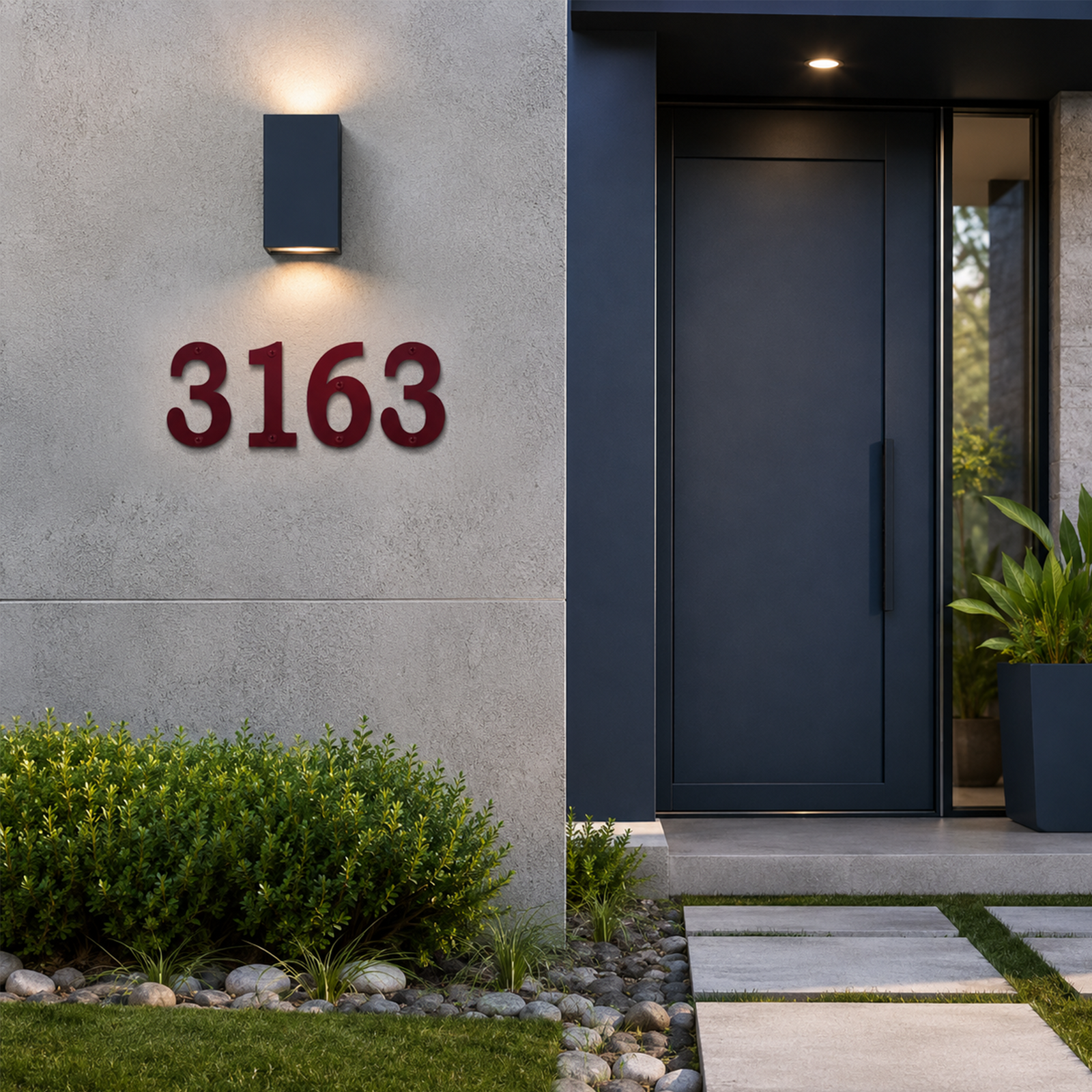Deep burgundy red metal house numbers featuring the digits "3163" are installed on a smooth gray concrete facade. The setting features a modern navy blue entryway door with a long vertical handle, a lit rectangular wall sconce, and a lush green hedge lining a stone paver path.