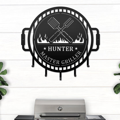 Grill Master Name Sign with Metal Hooks featuring a black circular design with crossed spatulas and fire logo, ideal for organizing grilling utensils in outdoor kitchens or man caves.