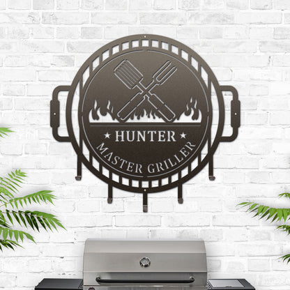 The Grill Master Name Sign with Metal Hooks showcases a metal sign featuring a grill logo, perfect for organizing utensils in outdoor kitchens, reflecting HouseSensationsArt's custom signage expertise.