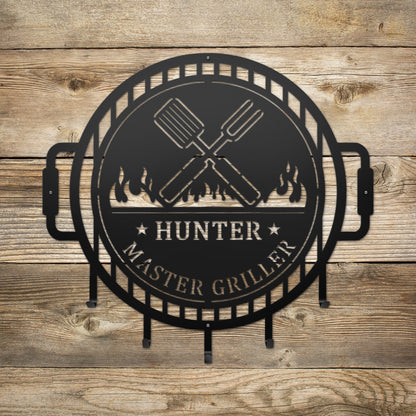 Grill Master Name Sign with Metal Hooks features a black metal sign with a grill logo and crossed spatulas, ideal for personalizing an outdoor kitchen with a stylish, organized touch.