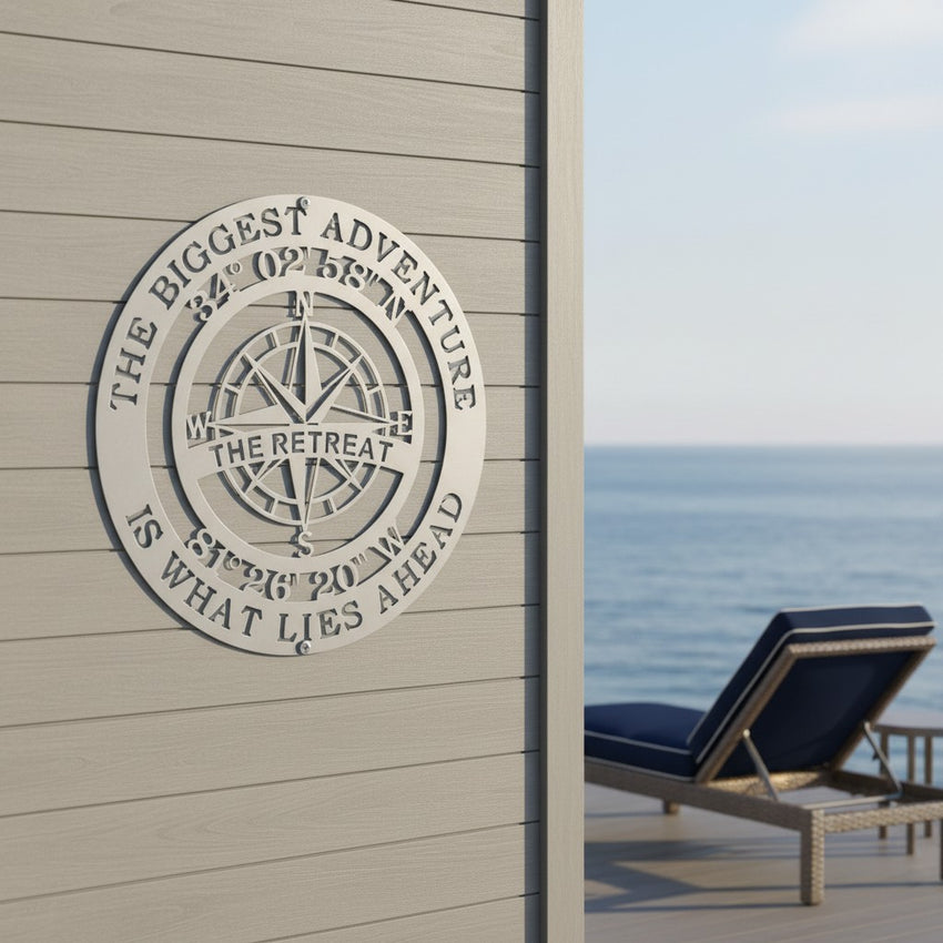 A metallic satin silver circular metal compass sign is installed on a light gray wooden exterior accent wall. The plaque details geographical coordinates and the name "THE RETREAT." A contemporary deck area with a modern lounge chair and a distant ocean view completes the setting.