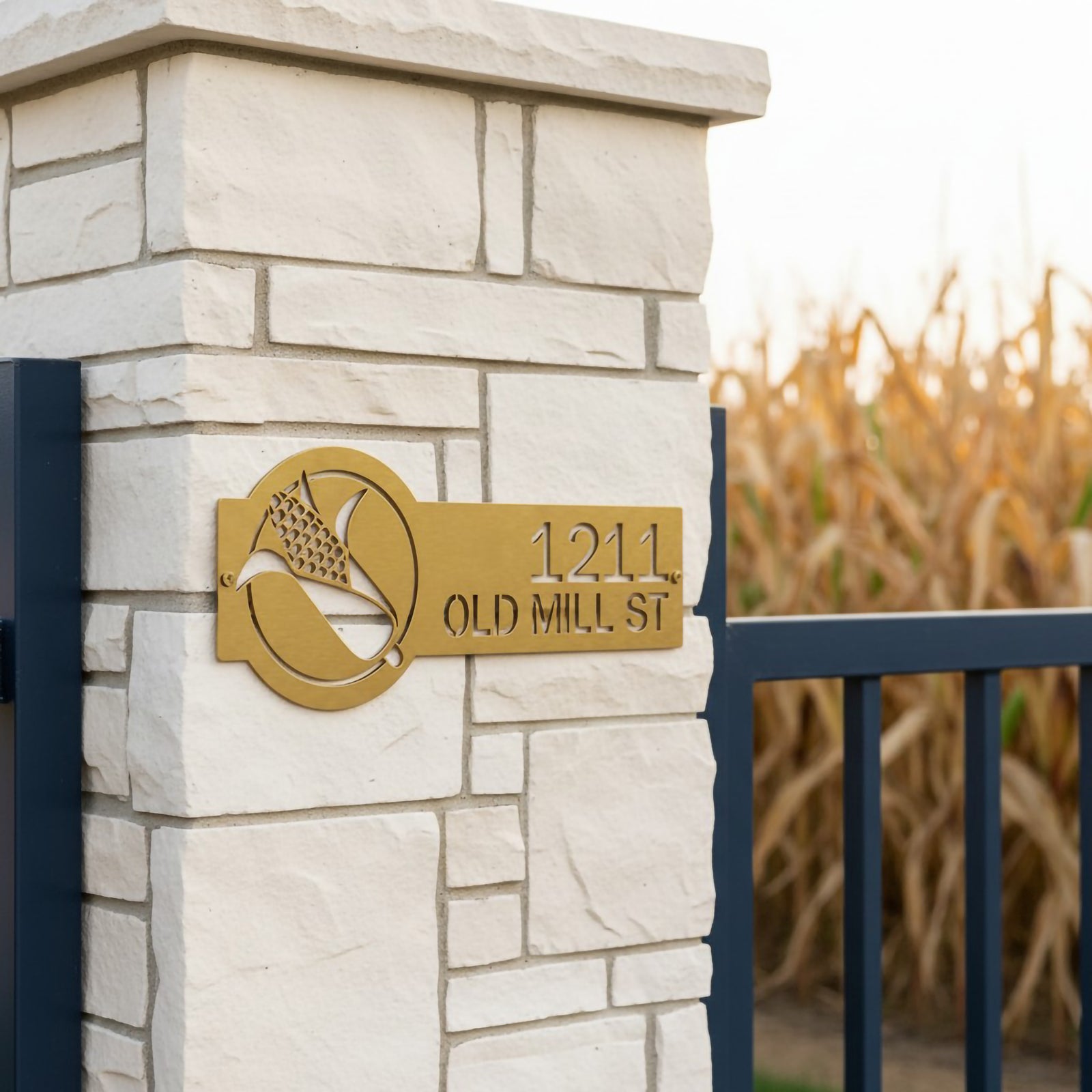 Metallic gold corn address sign with 1211 Old Mill St mounted on a white stone pillar beside a cornfield