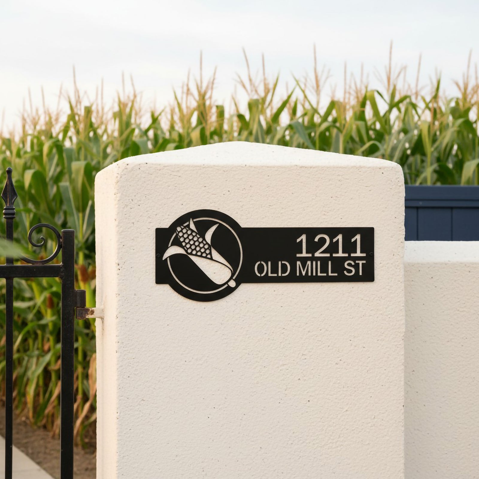 Metallic gold corn house number sign with 1211 Old Mill St mounted on a white stone pillar beside a farm field