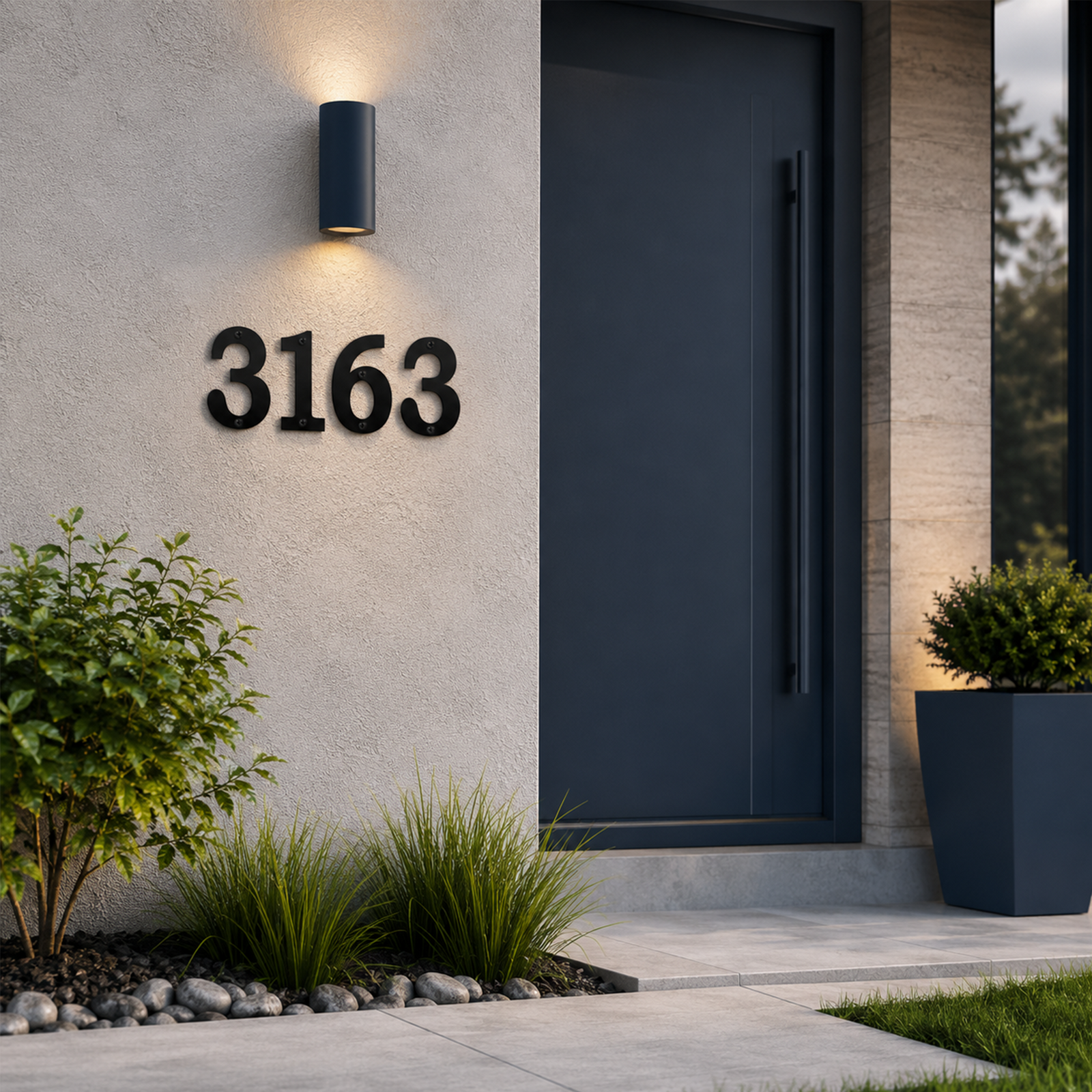 High-contrast satin black house numbers displaying "3163" are mounted on a light textured exterior wall next to a large dark gray modern front door. A sleek black sconce shines downward onto the numbers, surrounded by minimalist landscaping with green ornamental grasses and smooth river rocks.
