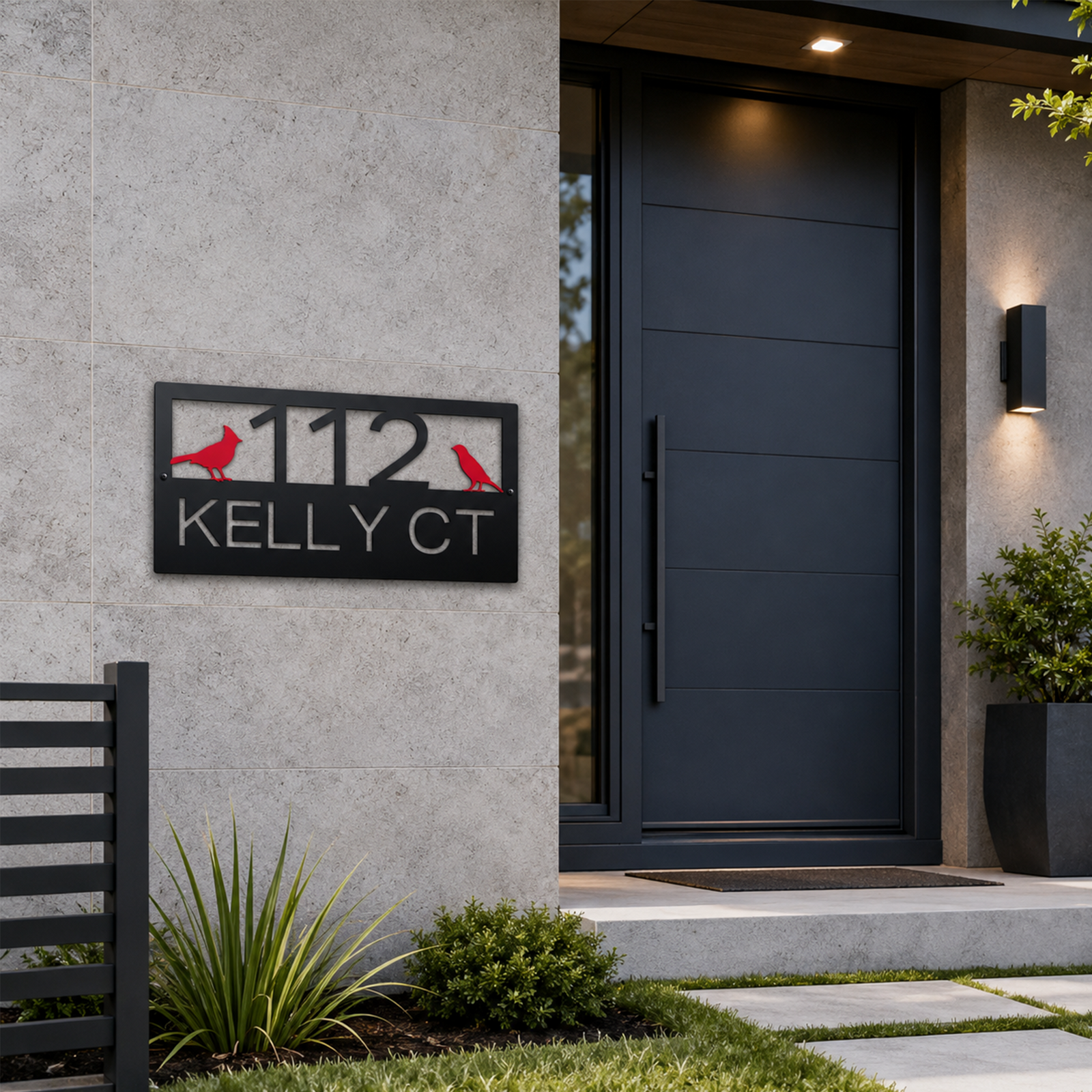 A satin black rectangular metal plaque reading "112 KELLY CT" with two red cardinal silhouettes is mounted next to a minimalist dark gray front door. The clean exterior features stone tiles, integrated lighting, a sleek vertical door handle, and simple green potted foliage.
