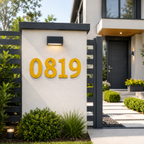 Bright golden yellow house numbers "0819" mounted on a textured white concrete fence pillar beneath a black minimalist outdoor light fixture, surrounded by modern landscaping.