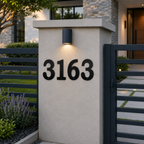Large, black serif metal house numbers reading "3163" are mounted horizontally on a light gray stucco entryway pillar. A cylindrical black downlight illuminates the numbers from above, flanked by a dark metal fence and purple lavender bushes in a manicured modern yard.