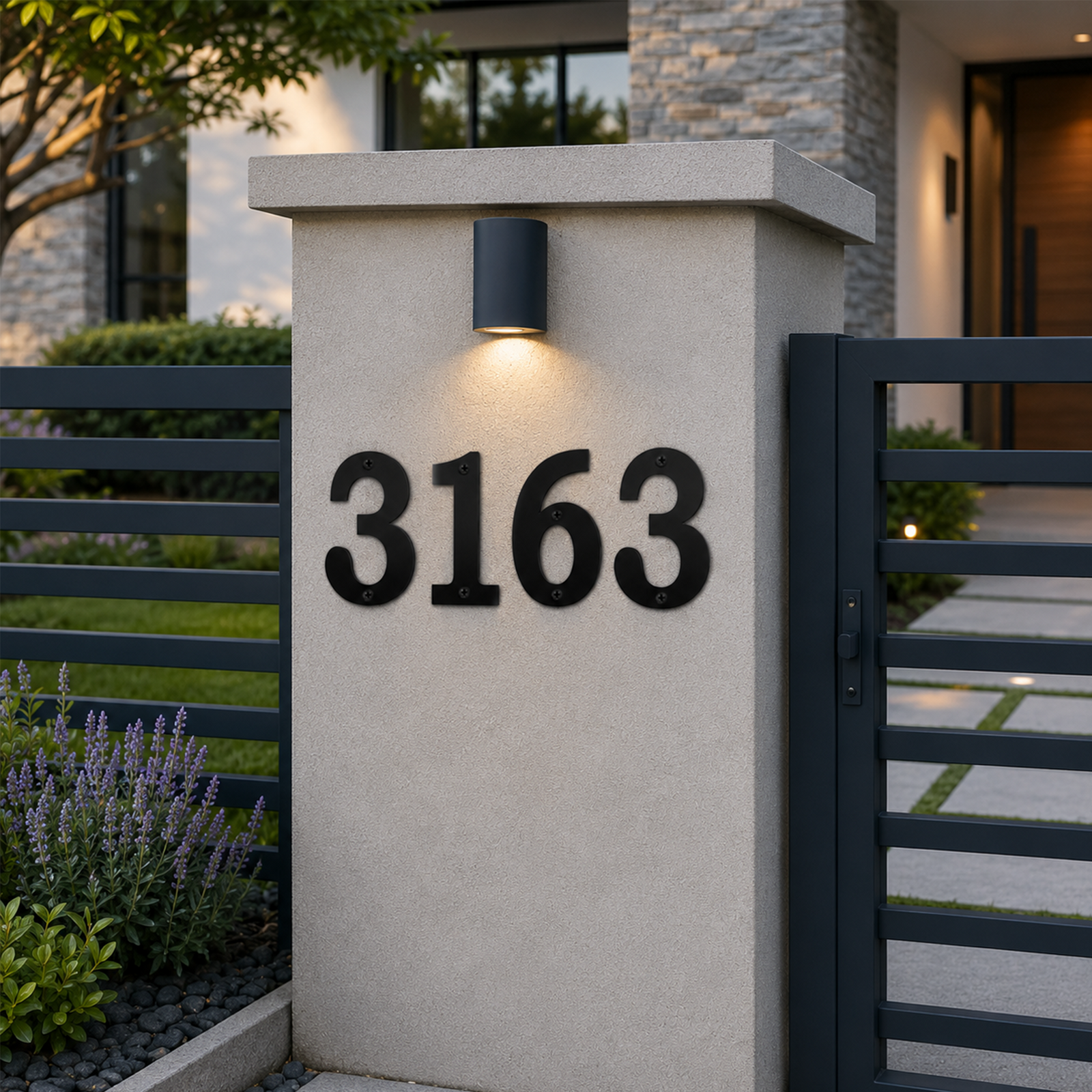 Large, black serif metal house numbers reading "3163" are mounted horizontally on a light gray stucco entryway pillar. A cylindrical black downlight illuminates the numbers from above, flanked by a dark metal fence and purple lavender bushes in a manicured modern yard.