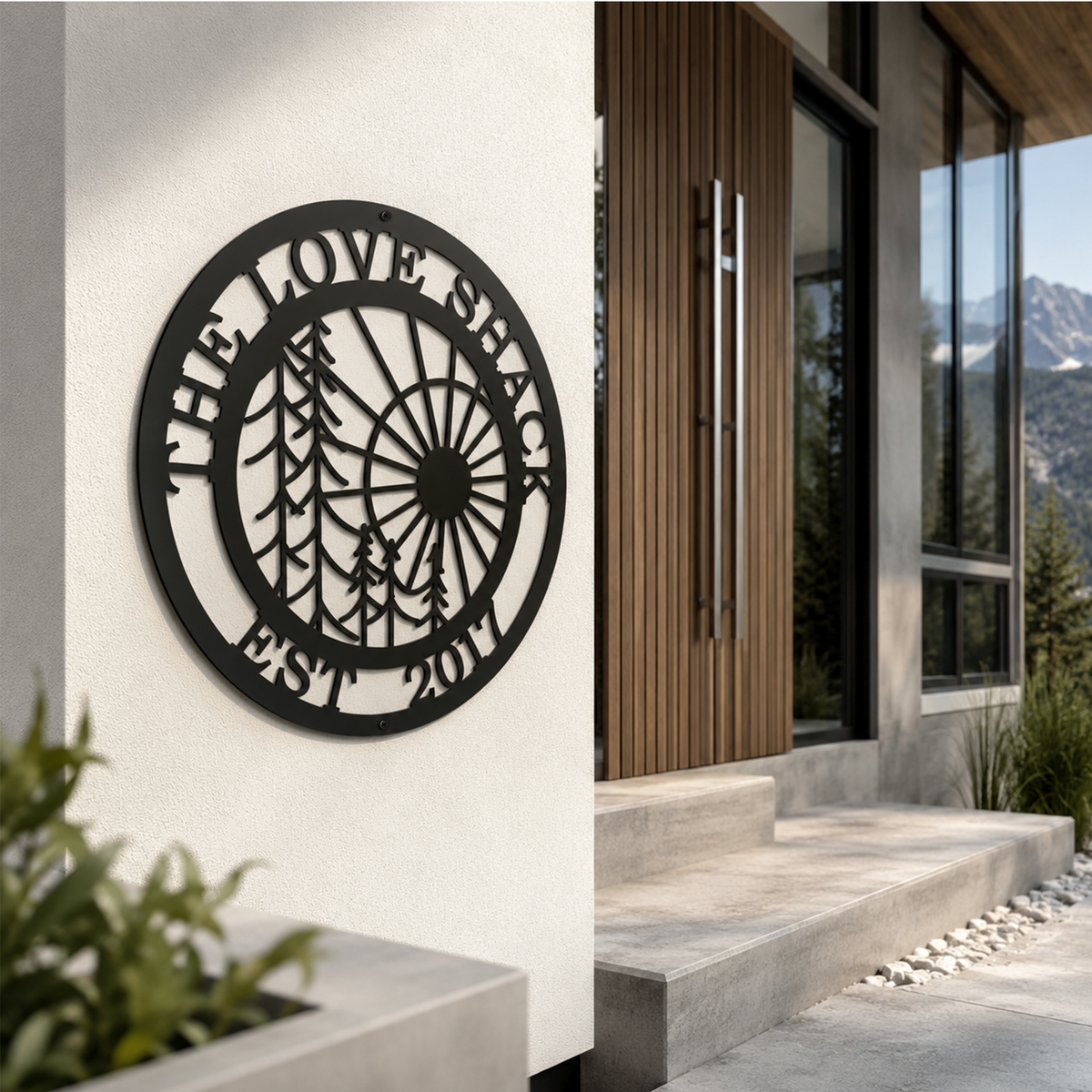 Black custom family metal wall sign with forest and sun design displayed on a modern outdoor stucco wall beside a wooden front door