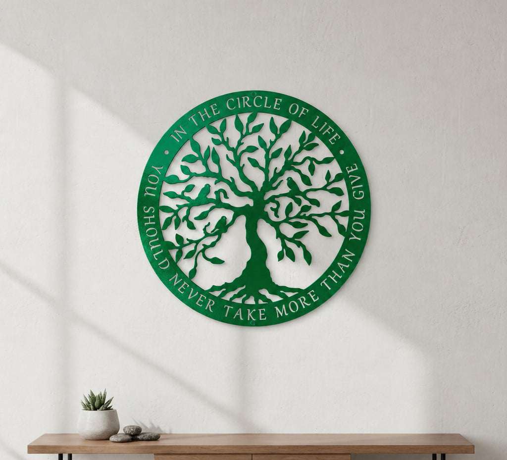 A circular emerald green metal sign featuring a laser-cut Tree of Life and an inspirational quote about the circle of life running along the circular border. The sign is mounted on a plain white textured wall above a clean wooden table holding a small potted succulent.