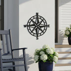 A matte black metal compass rose sign mounted on a white porch wall, positioned behind a dark grey rocking chair and a blue ceramic pot filled with white hydrangeas.