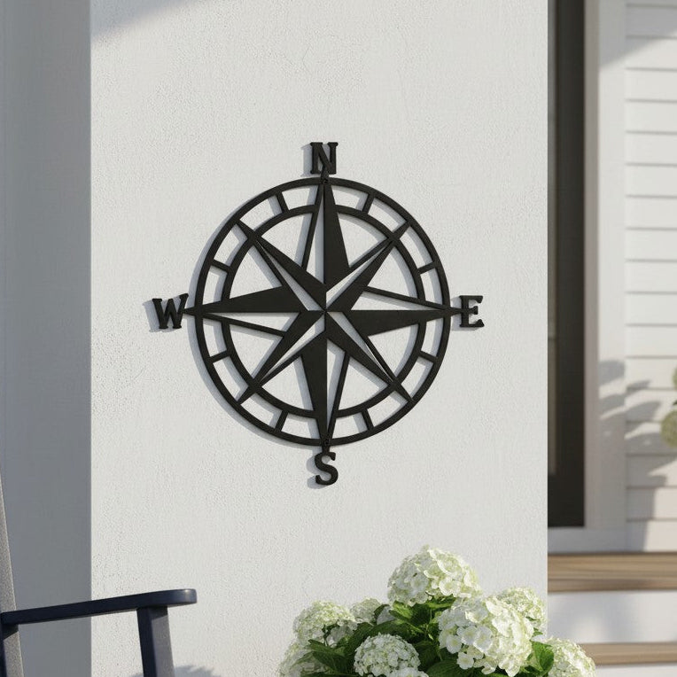 A matte black metal compass rose sign mounted on a white porch wall, positioned behind a dark grey rocking chair and a blue ceramic pot filled with white hydrangeas.