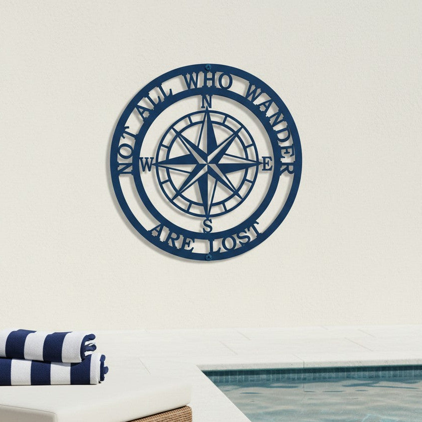 A round, vibrant blue metal wall plaque displaying a nautical compass and the phrase "NOT ALL WHO WANDER ARE LOST". The sign is anchored to a plain off-white exterior wall above a luxury swimming pool deck featuring a wicker lounger with rolled blue-and-white striped towels.