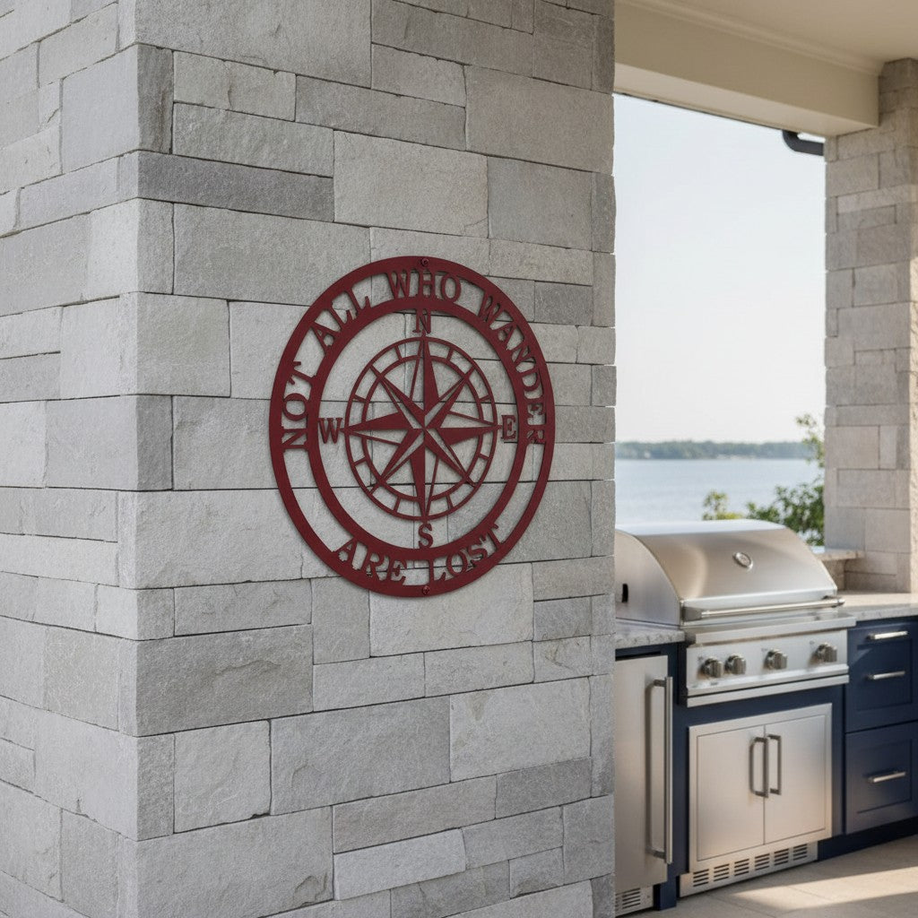 A deep crimson red metal compass sign inscribed with the quote "NOT ALL WHO WANDER ARE LOST". It is mounted to a gray stone-brick column in a modern outdoor kitchen area equipped with a stainless steel grill setup and a distant view of the water.