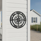 A circular matte black steel compass plaque featuring the quote "NOT ALL WHO WANDER ARE LOST". The piece is mounted on the clean white siding of a coastal home pillar, with a sandy beach path and a blurred white cottage in the background.