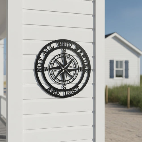 A circular matte black steel compass plaque featuring the quote "NOT ALL WHO WANDER ARE LOST". The piece is mounted on the clean white siding of a coastal home pillar, with a sandy beach path and a blurred white cottage in the background.