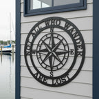 A close-up of a circular matte black metal cut-out sign depicting a classic compass design with the text "NOT ALL WHO WANDER ARE LOST". The plaque is installed on light gray horizontal vinyl siding, with a marina, water, and blurred sailboats visible in the background.