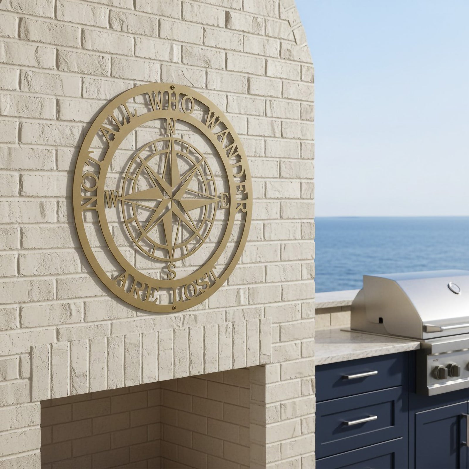 A round custom metal sign in a brushed gold finish, featuring a detailed geometric compass rose surrounded by the words "NOT ALL WHO WANDER ARE LOST". The sign is centered on a painted white brick outdoor fireplace mantel overlooking a calm blue ocean.