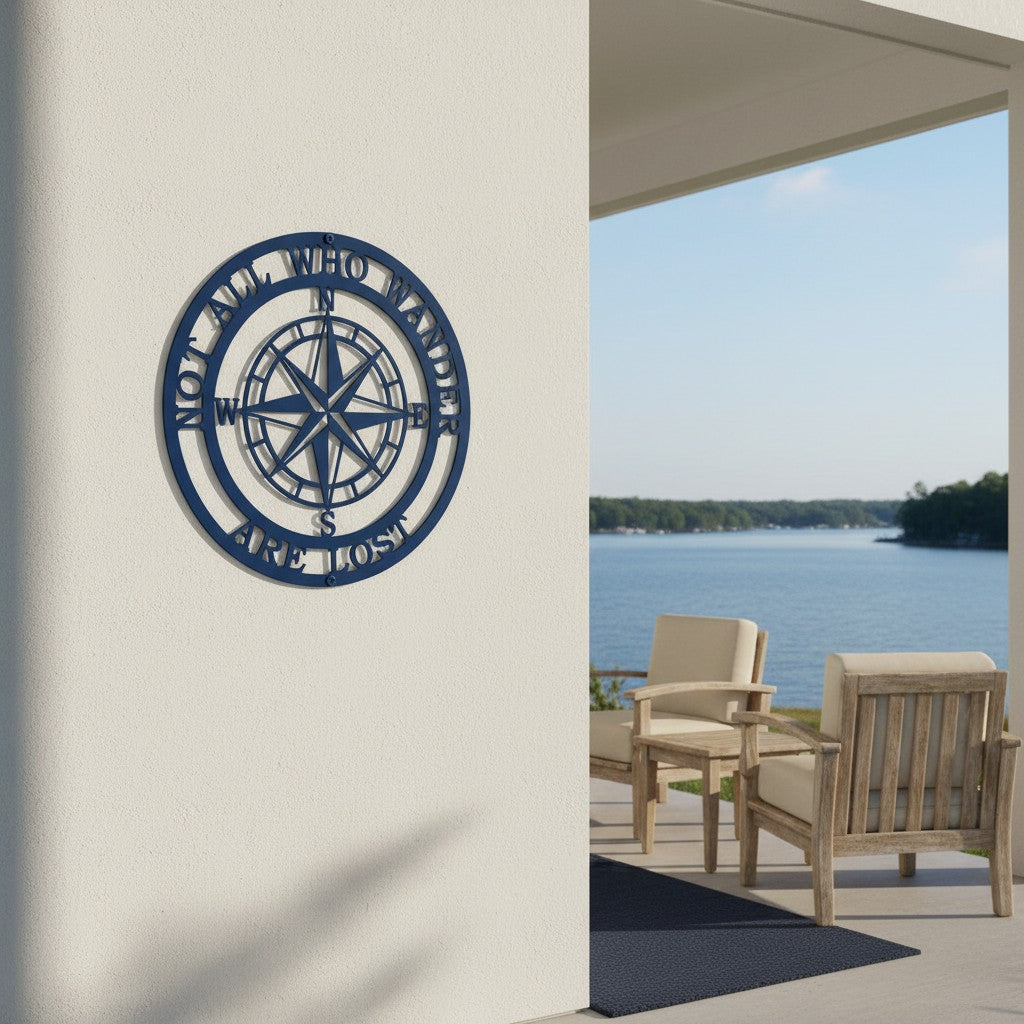 A round, laser-cut navy blue metal wall sign featuring an intricate compass rose in the center. The circular border contains the phrase "NOT ALL WHO WANDER ARE LOST". The sign is mounted on a smooth cream stucco pillar next to an outdoor covered patio with wooden lounge chairs overlooking a serene lake.