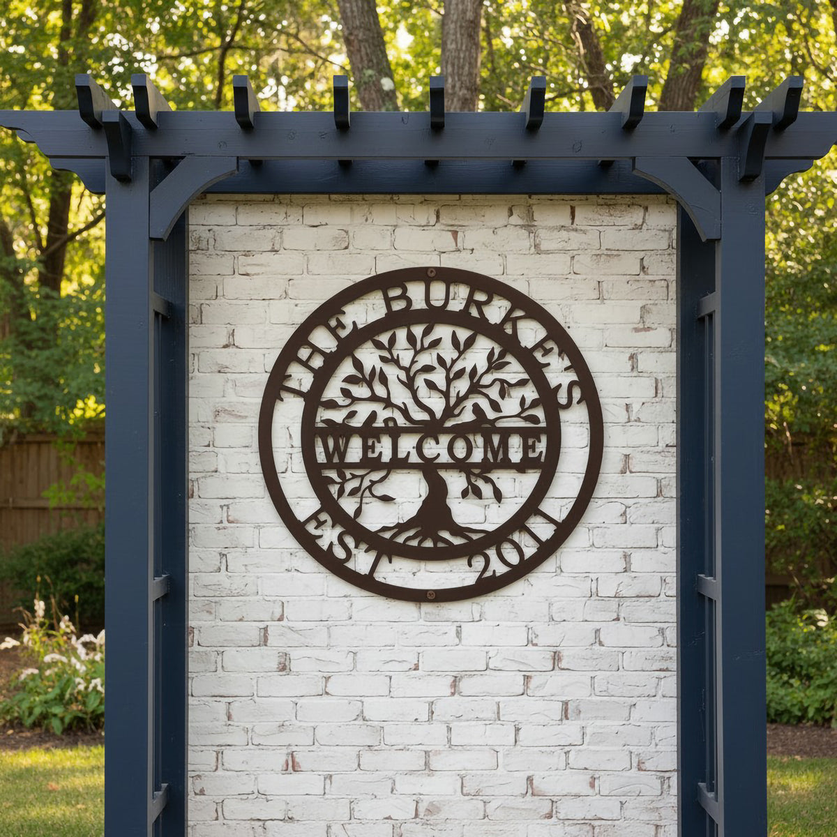A dark brown circular metal sign personalized for "The Burkes Welcome Est. 2011," displayed on a white brick structural wall beneath a navy blue garden arbor structure.