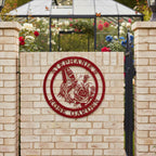 A deep red round metal sign personalized with "Stephanie's Rose Garden," mounted on a light beige brick wall in front of a glass greenhouse filled with blooming roses.