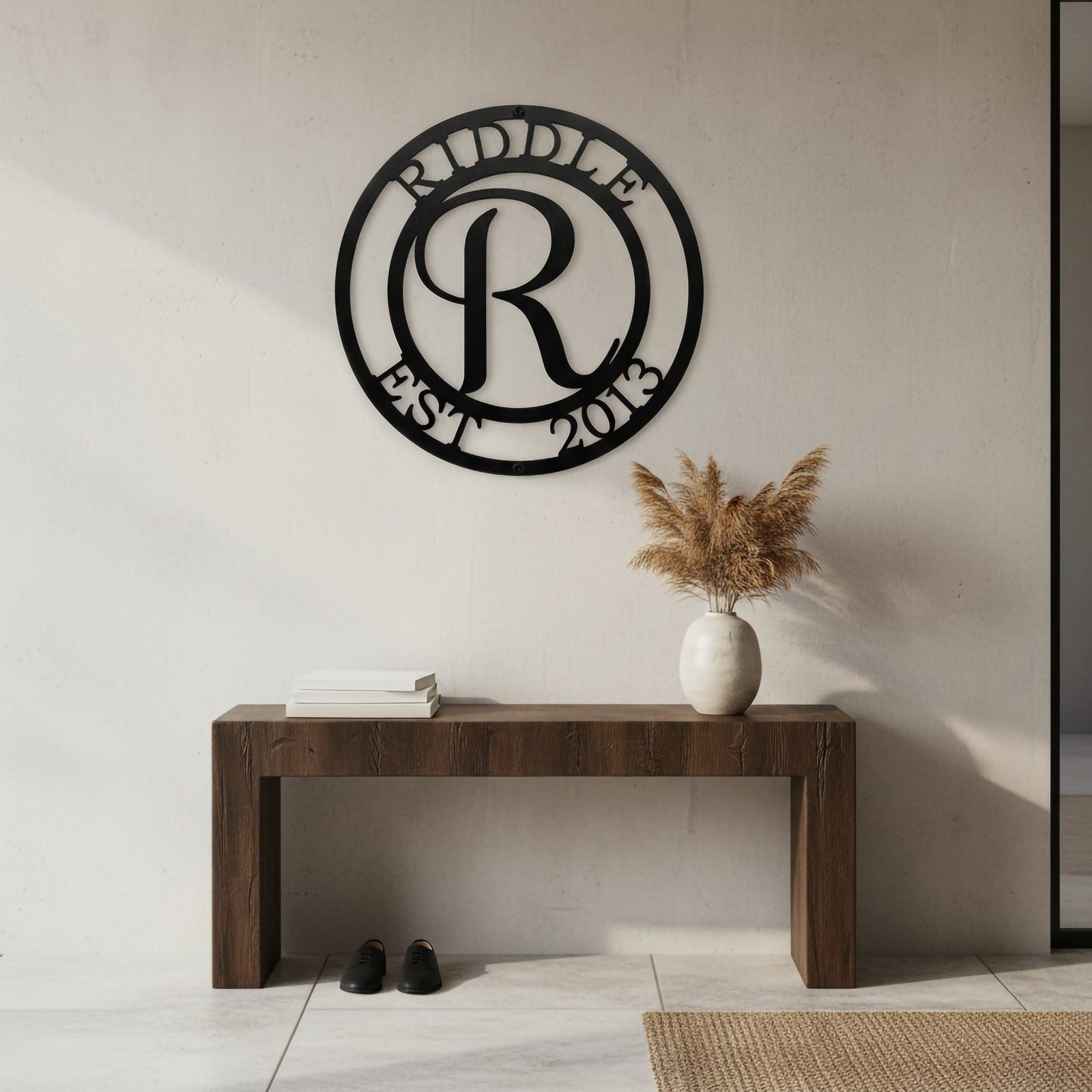 Satin black personalized circle monogram metal sign with "RIDDLE" and "EST 2013" cutout text mounted on a plain white wall above a wooden entryway table with dried grasses.
