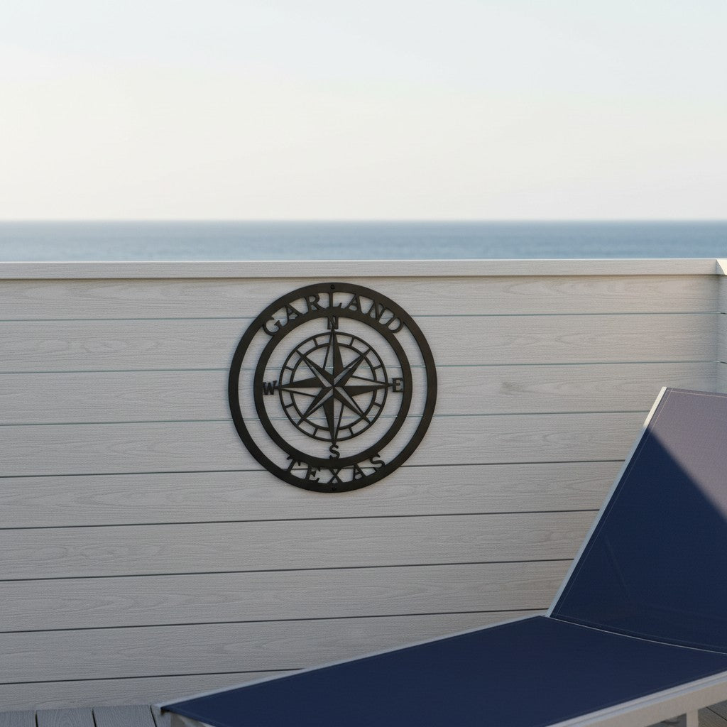  A black round metal compass rose sign reading "Garland Texas" along the outer ring, mounted on a white horizontal wood privacy wall on an oceanfront deck next to a blue lounge chair.