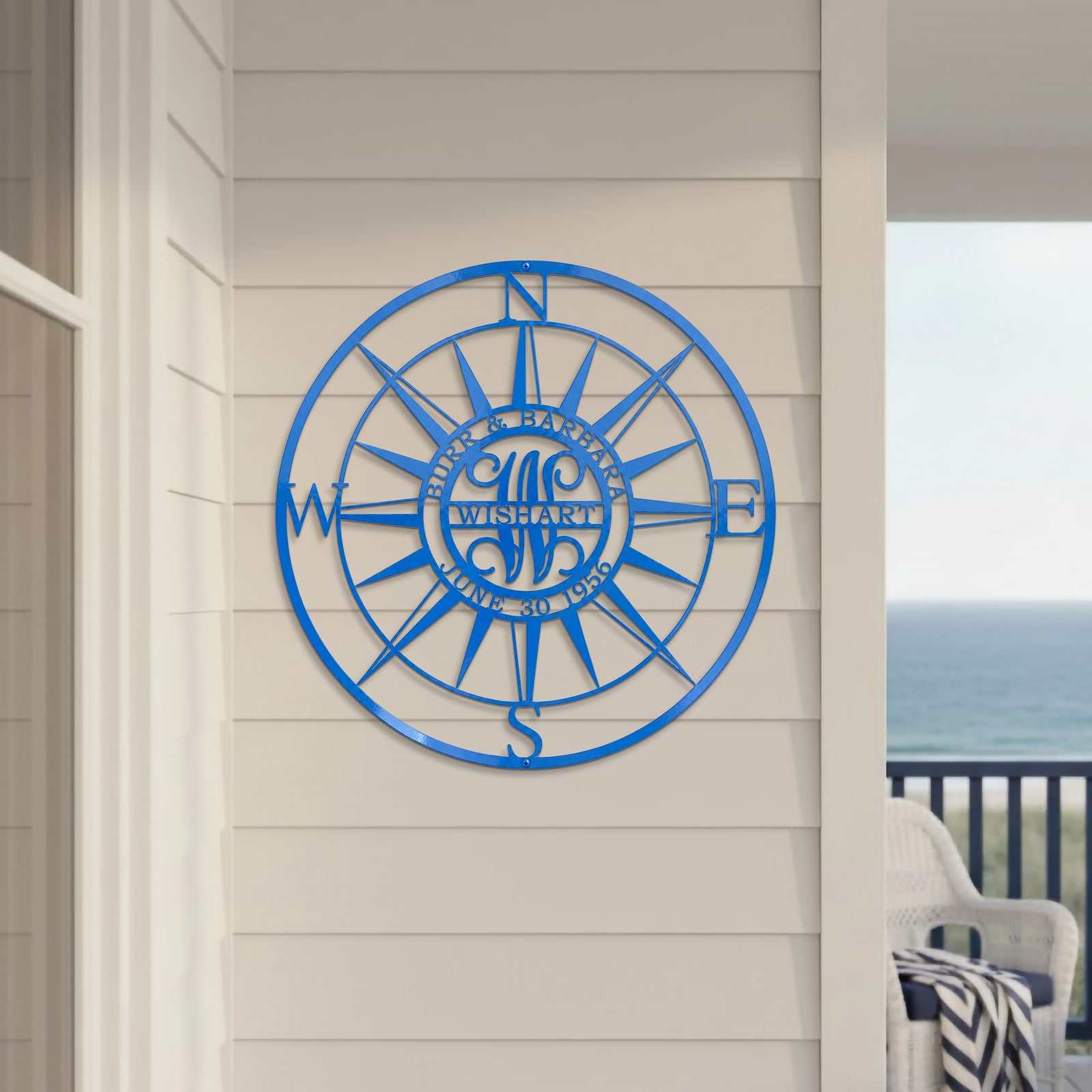 A round custom metal sign in a vibrant royal blue finish, showcasing a detailed compass rose configuration personalized with the name "WISHART". The sign is fixed to a light tan wall next to an open balcony featuring a white wicker chair and a wide view of the blue sea.