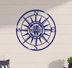 A round, laser-cut deep royal blue metal compass rose sign mounted centered on a light-colored horizontal panel wall. The middle ring of the compass features the personalized cutout lettering "BURR & BARBARA" on top, "JUNE 30 1956" on the bottom, and a stylized "WISHART" monogram in the center.