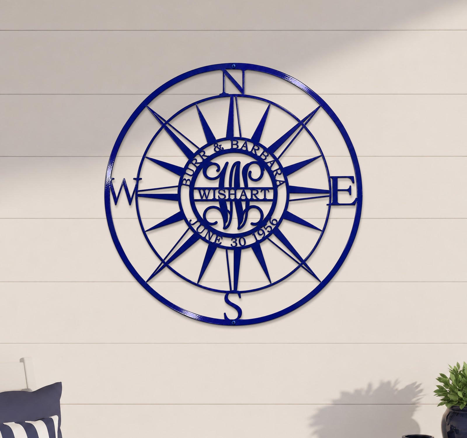 A round, laser-cut deep royal blue metal compass rose sign mounted centered on a light-colored horizontal panel wall. The middle ring of the compass features the personalized cutout lettering "BURR & BARBARA" on top, "JUNE 30 1956" on the bottom, and a stylized "WISHART" monogram in the center.