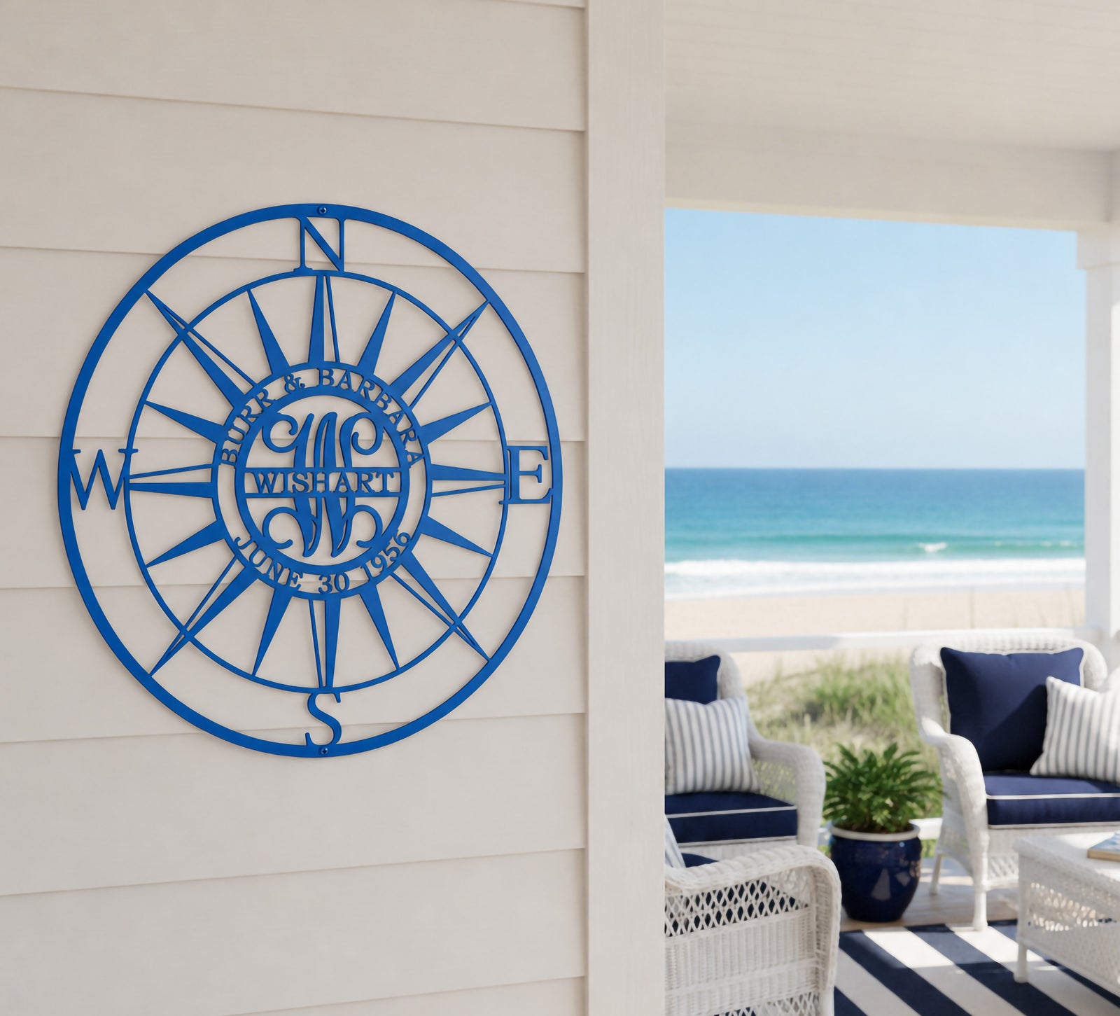 A circular panther blue metal outdoor compass rose sign featuring a custom "WISHART" wedding anniversary monogram. The piece is installed on white horizontal lap siding on a coastal porch, with white wicker patio chairs, a striped rug, and a clear blue ocean visible in the background.