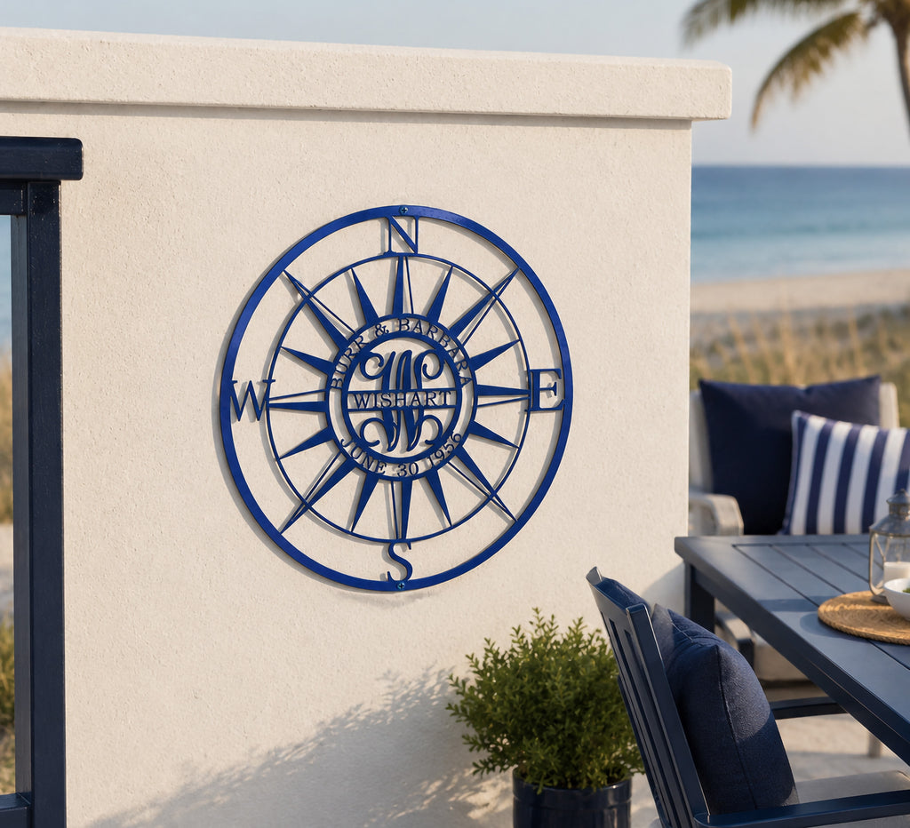 A customized dark blue metal compass sign inscribed with "BURR & BARBARA WISHART JUNE 30 1956". It is securely mounted on a low, white-painted stucco deck wall alongside an outdoor patio dining area right next to a sandy beach and turquoise ocean water.