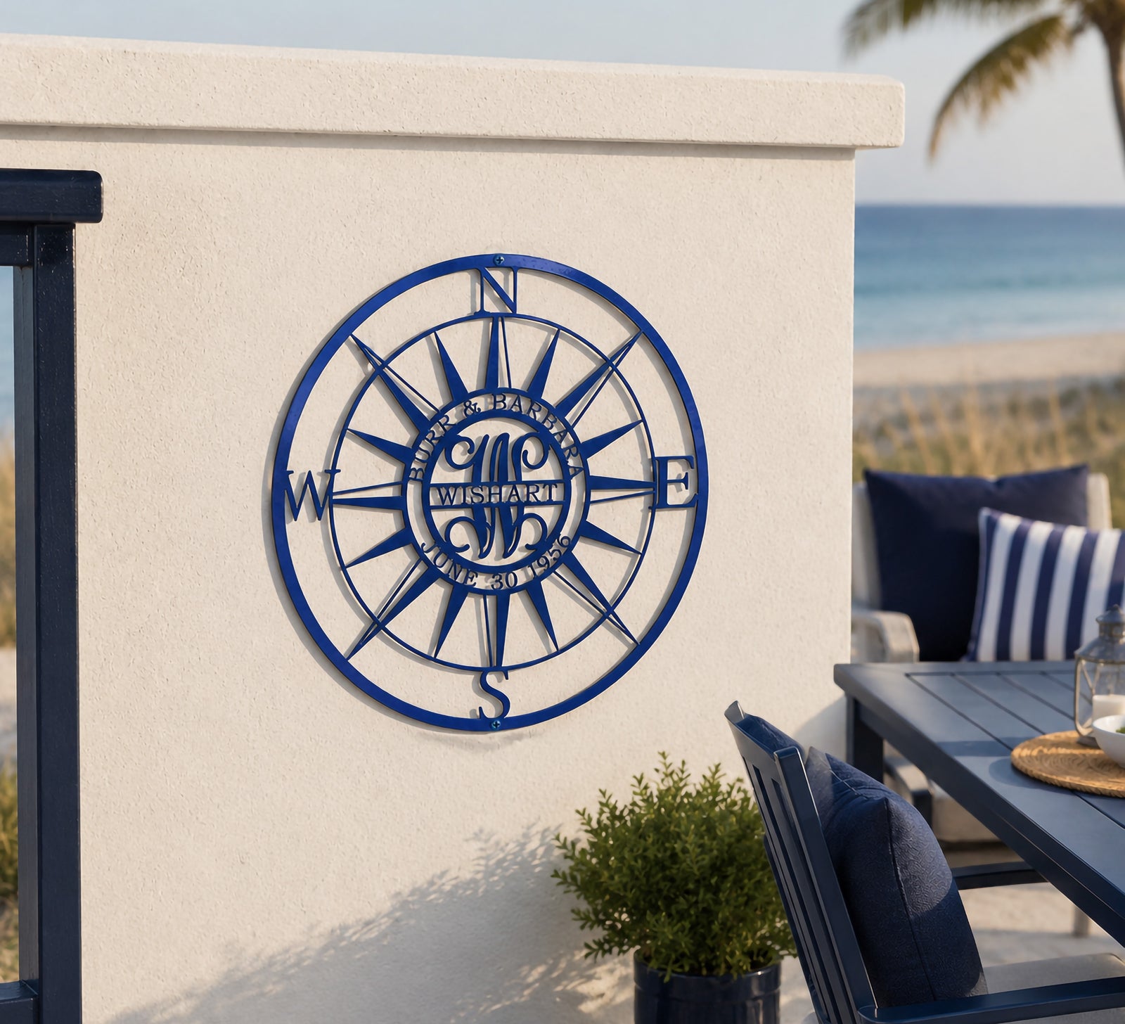 A customized dark blue metal compass sign inscribed with "BURR & BARBARA WISHART JUNE 30 1956". It is securely mounted on a low, white-painted stucco deck wall alongside an outdoor patio dining area right next to a sandy beach and turquoise ocean water.