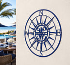 An angled view of a circular royal blue metal family name compass plaque displaying the name "WISHART" and the date "JUNE 30 1956". It is mounted to a smooth white exterior column next to a luxury patio dining table set, overlooking a swimming pool, palm trees, and the ocean beach.