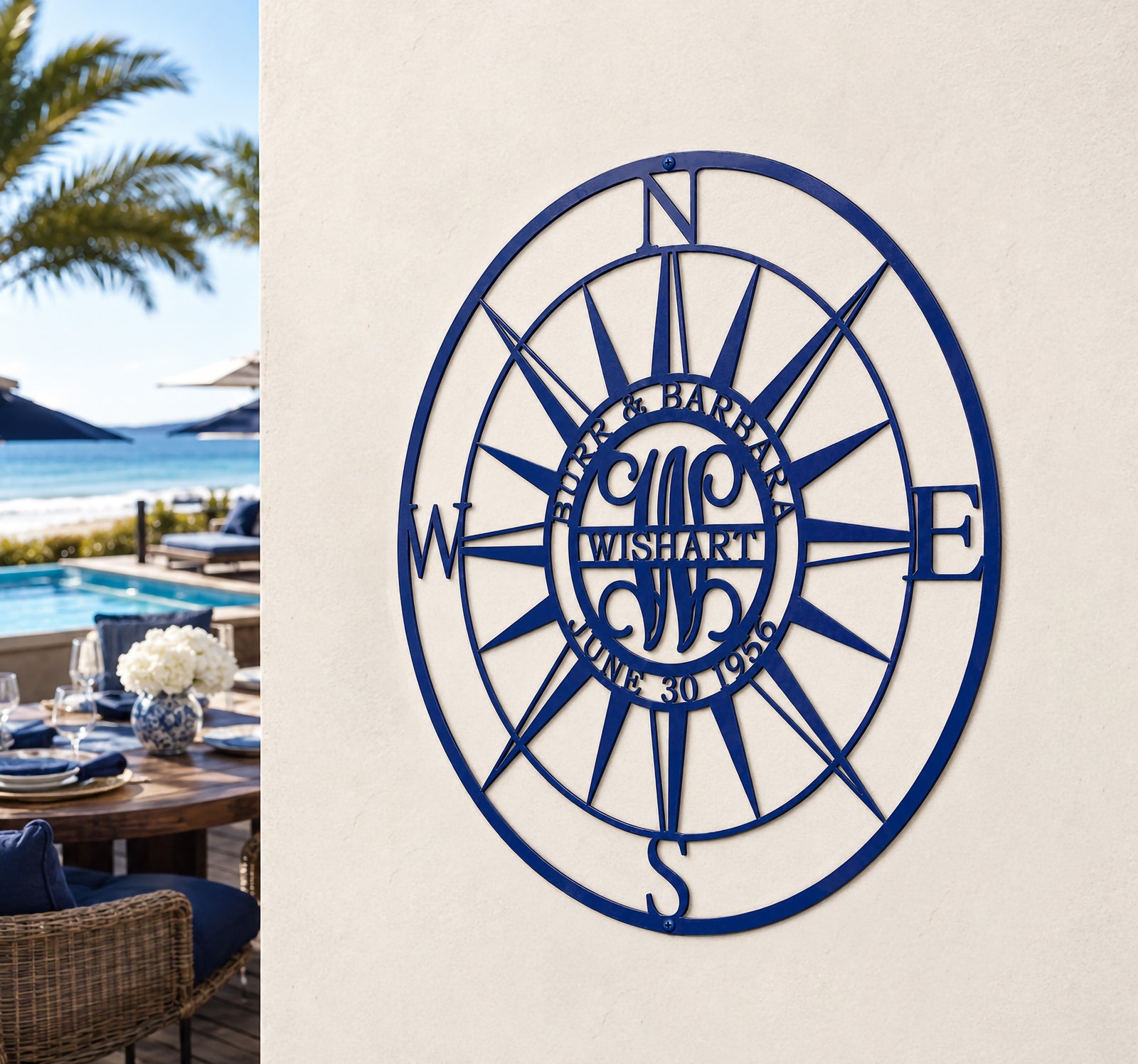 An angled view of a circular royal blue metal family name compass plaque displaying the name "WISHART" and the date "JUNE 30 1956". It is mounted to a smooth white exterior column next to a luxury patio dining table set, overlooking a swimming pool, palm trees, and the ocean beach.