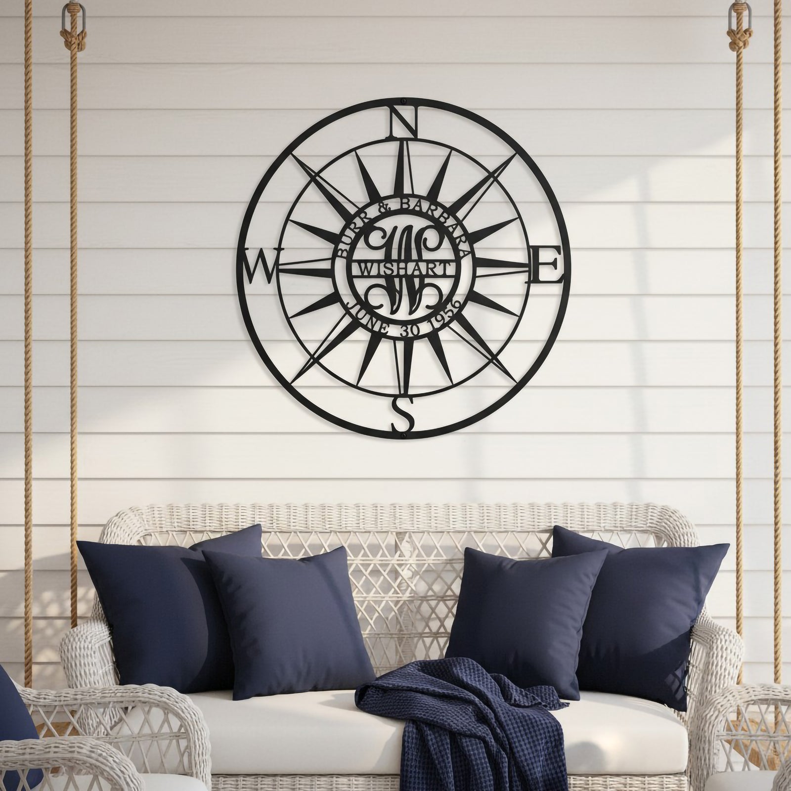 A round satin black steel family monogram sign featuring a classic nautical compass design customized with "WISHART EST. JUNE 30 1956". The piece is mounted on a clean white vertical plank wall directly above a white wicker hanging porch swing styled with dark blue throw pillows and a blanket.