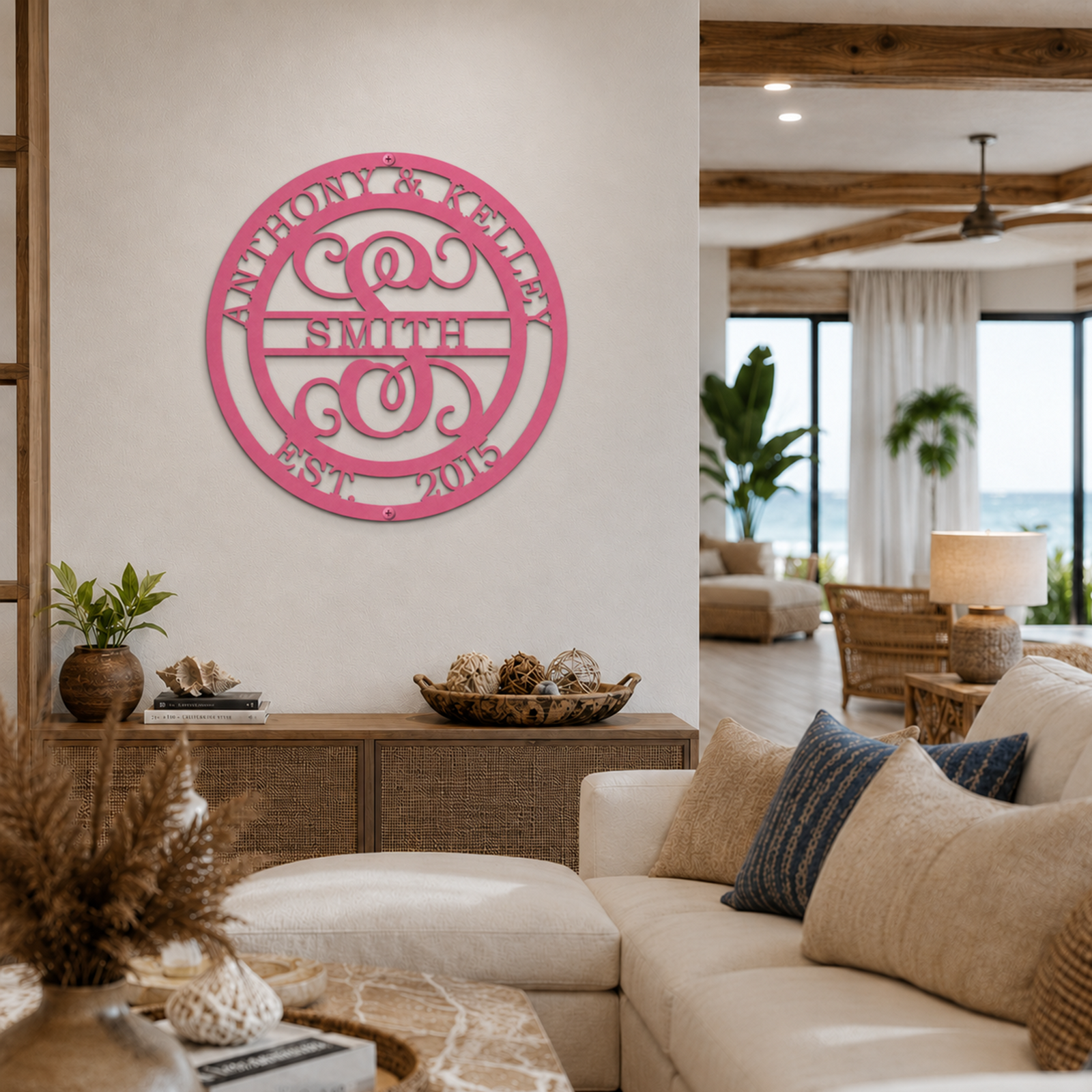 A circular metal monogram sign finished in a vibrant coral pink color is mounted on a white living room wall. The laser-cut sign features a large stylized initial "S" in the center with the name "SMITH" across a middle banner, flanked by "ANTHONY & KELLEY" on the top arch and "EST. 2015" on the bottom. The background shows a coastal-themed living room with a white sectional sofa and a beach view.