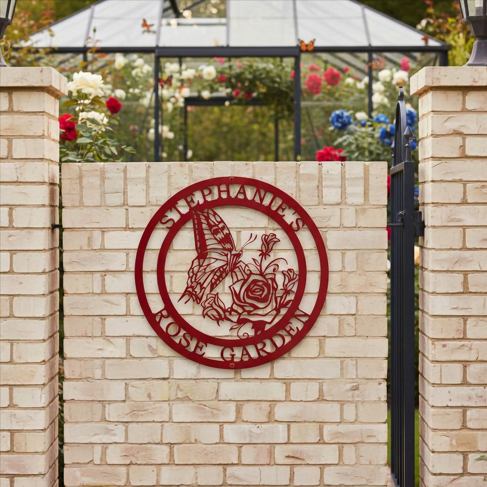 Matte desert crimson finish of the custom circular butterfly rose sign with text Stephanie's Rose Garden, mounted flat on a white brick courtyard wall with a glass-paneled garden greenhouse in the background.