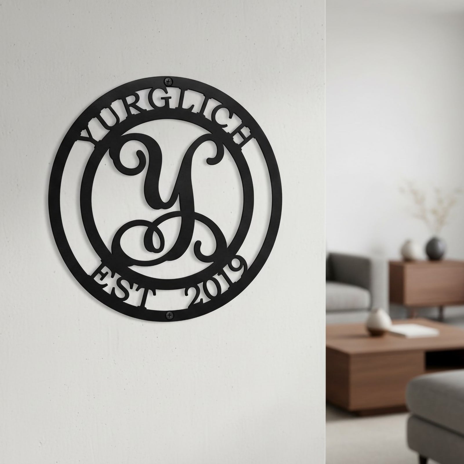 Satin black custom circular metal family name sign with a cursive initial monogram hanging in a clean, modern minimalist living room setting.