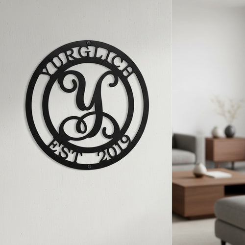 Satin black custom circular metal family name sign with a cursive initial monogram hanging in a clean, modern minimalist living room setting.