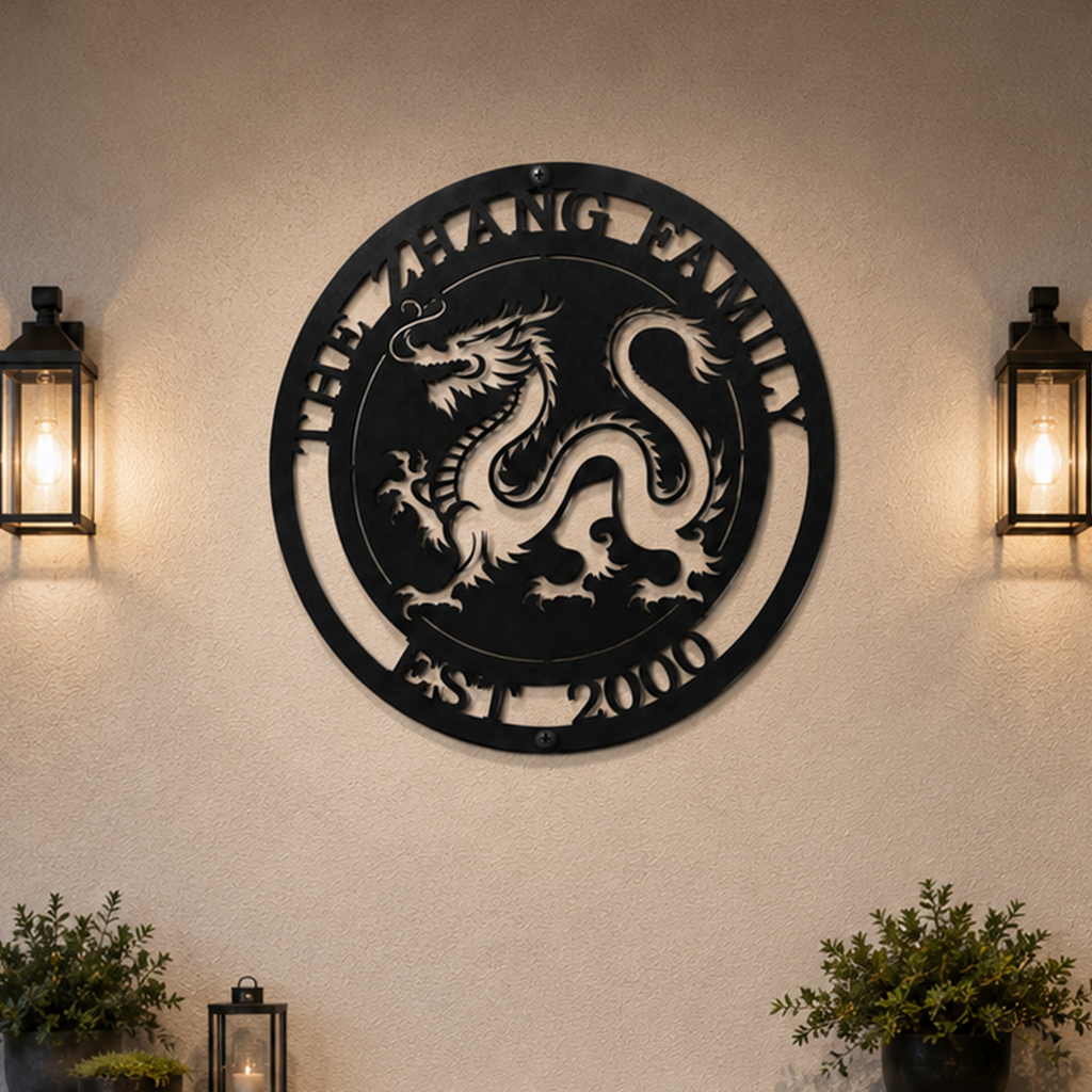 A textured charcoal-colored circular metal sign displaying an Imperial Chinese dragon with custom family text. The sign is mounted on a cream plaster wall, symmetrically framed by two glowing black metal outdoor candle lanterns and low potted green bushes below.