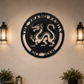A textured charcoal-colored circular metal sign displaying an Imperial Chinese dragon with custom family text. The sign is mounted on a cream plaster wall, symmetrically framed by two glowing black metal outdoor candle lanterns and low potted green bushes below.