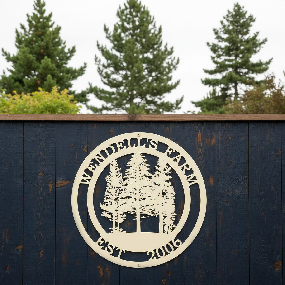 An off-white round metal sign personalized with "Wendell's Farm Est. 2006" featuring a cluster of pine trees, hanging on a dark navy blue wooden privacy fence.