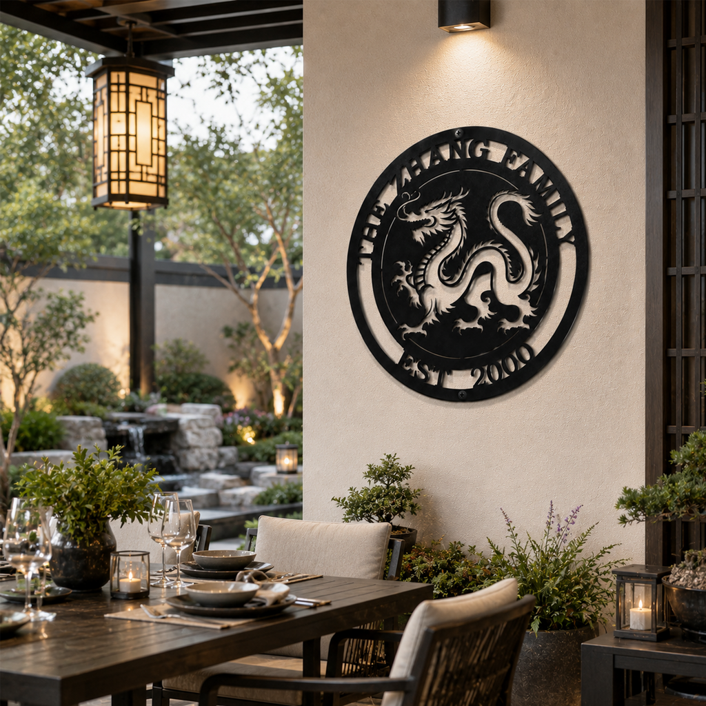 A circular metal medallion in a satin black finish displaying a detailed Chinese dragon cutout and the text "THE ZHANG FAMILY EST. 2000." The sign is installed on a modern cream pillar next to an elegant courtyard patio dining area, featuring a small waterfall fountain and stone landscaping in the background.