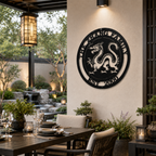 A circular metal medallion in a satin black finish displaying a detailed Chinese dragon cutout and the text "THE ZHANG FAMILY EST. 2000." The sign is installed on a modern cream pillar next to an elegant courtyard patio dining area, featuring a small waterfall fountain and stone landscaping in the background.