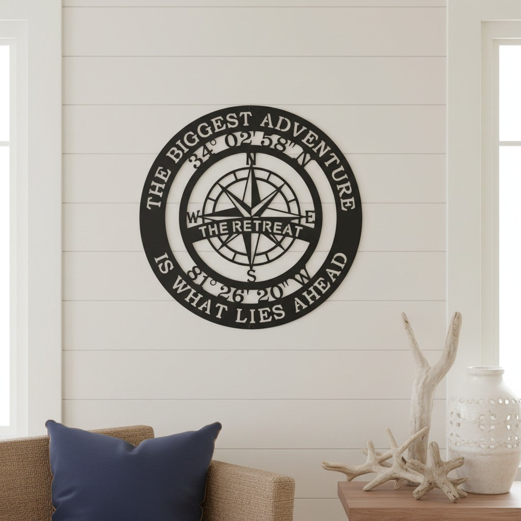 A matte black circular metal compass sign is mounted indoors on a white horizontal shiplap wall. The sign features custom geographic numbers and "THE RETREAT" cut out across the center. Below the sign sits a tan armchair with a blue throw pillow next to a side table adorned with white ceramic vases and decorative bleached driftwood branches.