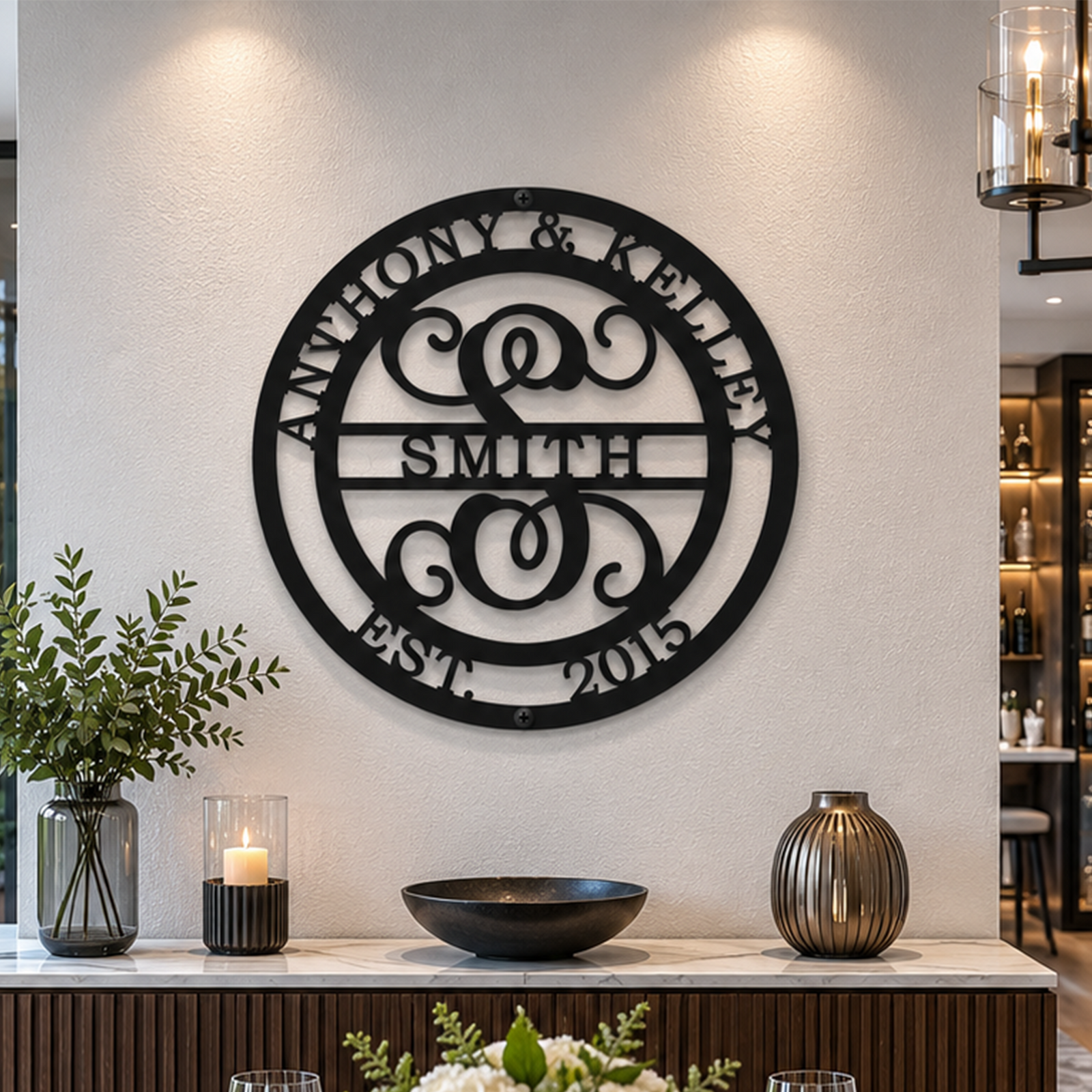 A classic matte black circular metal monogram plaque featuring the name "SMITH" is mounted on a white accent wall under recessed spotlighting. The sign is positioned directly above a luxury marble-top wooden buffet cabinet arranged with a lit candle, a dark decorative bowl, and a metallic vase.