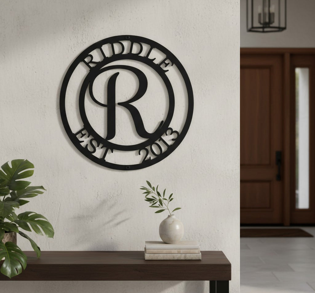 Satin black custom circular monogram metal plaque featuring the initial letter R, displayed on an indoor white stucco partition wall above a chunky rustic wooden bench table holding pampas grass.